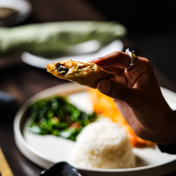 A hand holding a crispy spring roll, revealing its savory filling, with rice and greens in the background.