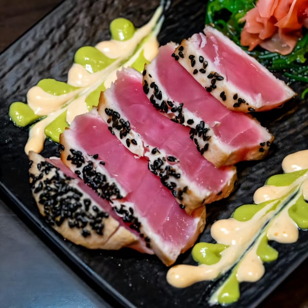 Seared tuna slices with black sesame seeds, green and orange sauces, seaweed, and pickled ginger.